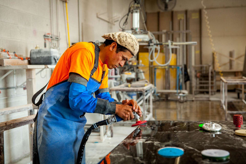 Stone fabricator polishing dark granite slab with pneumatic wet polisher and diamond pad in fabrication shop